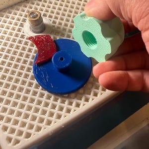 Op de afbeelding: Een blauw cirkelvormig object met een rood, gebogen stuk en een lichtgroen, tandwielvormig object met een schroefdraadgat. De objecten bevinden zich op een wit rooster. Een hand houdt het groene object vast.