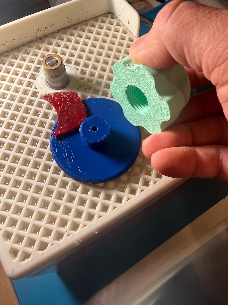 Op de afbeelding: Een blauw cirkelvormig object met een rood, gebogen stuk en een lichtgroen, tandwielvormig object met een schroefdraadgat. De objecten bevinden zich op een wit rooster. Een hand houdt het groene object vast.