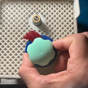 Op de afbeelding: Een hand die een lichtgroen, wit en blauw object vasthoudt. Het object heeft een cirkelvormig ontwerp met concentrische ringen. Een rood en blauw object is ook zichtbaar.