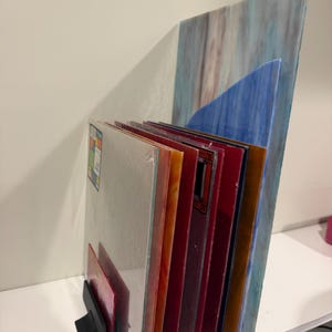 May include: A collection of rectangular stained glass sheets arranged vertically in a black metal stand. The glass pieces feature various colors including blue, red, orange, and amber, with some sheets displaying marbled textures and others showing solid hues.