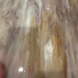 May include: A close-up of a piece of stained glass with a swirling pattern. The glass features a blend of colors, including shades of brown, white, and purple. The light reflects off the surface, creating a shimmering effect.