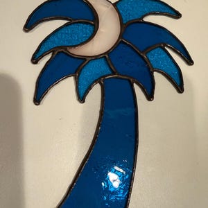 May include: Stained glass palm tree suncatcher with a crescent moon. The palm tree is made of shades of blue glass with a white moon. The suncatcher is framed with a dark metal border and hangs from a chain.