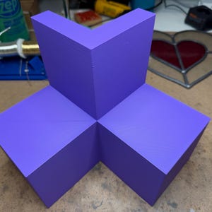 May include: A geometric, purple-painted wooden sculpture with a cross-like design. The sculpture is composed of three rectangular blocks joined at right angles, forming a three-dimensional shape. The color is a vibrant, solid purple.