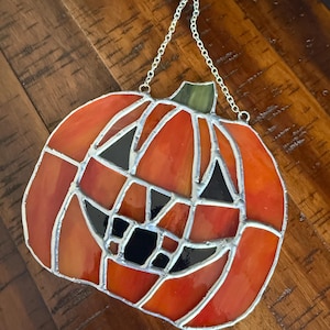 May include: Stained glass pumpkin decoration with a smiling face, featuring orange, red, and black glass pieces. The pumpkin has a green stem and is suspended by a silver chain, perfect for Halloween decor.