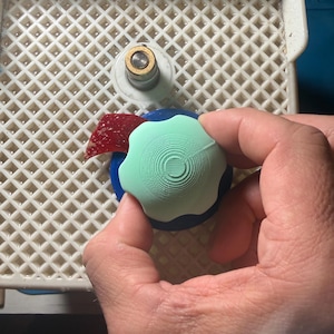 Op de afbeelding: Een lichtgroen, bloemvormig object met concentrische cirkeldetails wordt boven een blauwe basis gehouden. Een klein stukje rood glas en een wit en goudkleurig object zijn ook zichtbaar op een wit rooster.
