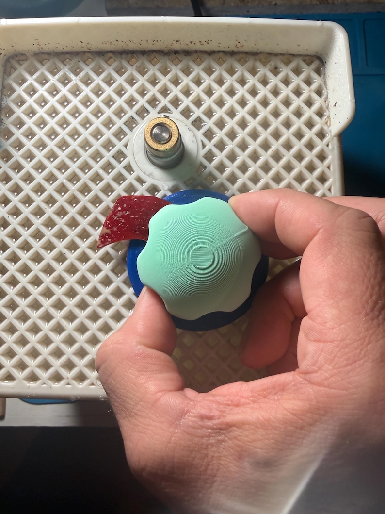 Op de afbeelding: Een lichtgroen, bloemvormig object met concentrische cirkeldetails wordt boven een blauwe basis gehouden. Een klein stukje rood glas en een wit en goudkleurig object zijn ook zichtbaar op een wit rooster.