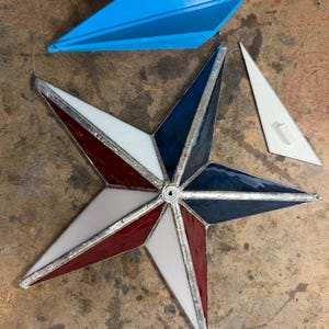 May include: A patriotic-themed stained glass star with red, white, and blue segments. The star is surrounded by a silver metal frame. A blue paper airplane and a white triangle are also present.