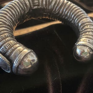 Puede incluir: Una pulsera de puño plateada con intrincados patrones y un extremo redondeado. La pulsera tiene un diseño acanalado a lo largo de su cuerpo curvo. Los extremos son lisos y reflectantes, con adornos detallados cerca del cierre.