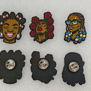 May include: Three enamel pins featuring illustrated portraits of individuals with diverse hairstyles and accessories. The top row shows colorful character designs, while the bottom row displays the black metal backs with circular pin fasteners.