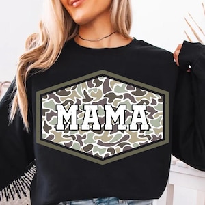 Może przedstawiać: Czarna bluza z długim rękawem z kamuflażem w kształcie sześciokąta. Słowo "MAMA" jest wydrukowane białymi literami blokowymi wewnątrz wzoru kamuflażu. Styl casual.