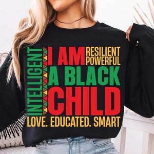 Op de afbeelding: Zwart sweatshirt met lange mouwen met de tekst "I AM A BLACK CHILD" in rood en groen. Extra tekst bevat "INTELLIGENT, RESILIENT, POWERFUL, LOVE, EDUCATED, SMART" in verschillende kleuren. Casual stijl.
