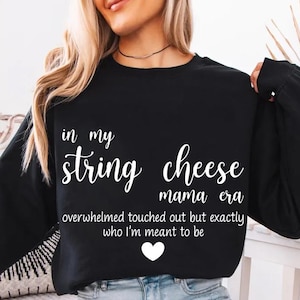 May include: Black sweatshirt with white text reading "in my string cheese mama era" and additional text below. A white heart is at the bottom. The sweatshirt is a casual style.