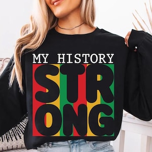 Puede incluir: Sudadera negra con el texto "MY HISTORY STRONG" en letras blancas y negras. La palabra "STRONG" está en un bloque rojo, verde y amarillo. La sudadera es de manga larga y la modelo lleva vaqueros azules.