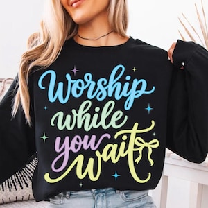 Peut inclure: Sweat-shirt noir avec l'inscription "Worship while you wait" en écriture colorée. Le texte est dans des tons de bleu, vert, violet et jaune. Le sweat-shirt a des manches longues et un col rond.