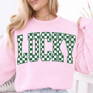 May include: A light pink sweatshirt with the word "LUCKY" in large, green and white checkered letters. The sweatshirt is long-sleeved and has a crew neck. The overall style is casual and comfortable.