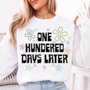 May include: White sweatshirt with the text "ONE HUNDERED DAYS LATER" in black, surrounded by colorful flower and circle graphics. The long-sleeved top is a casual style.