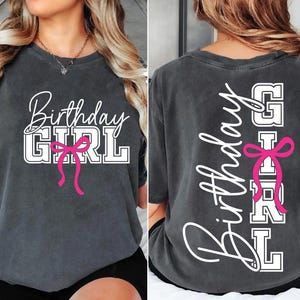 May include: Charcoal gray t-shirt with the words "Birthday GIRL" in white script and block letters, accented with a pink bow. The back of the shirt repeats the design vertically.