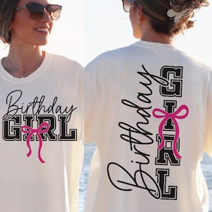 May include: Cream-colored t-shirt with the text "Birthday GIRL" in black script and block letters, accented with a pink bow. The back of the shirt repeats the text vertically. A casual, festive garment.