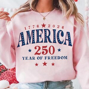 Puede incluir: Una sudadera rosa claro con el texto "1776 2026 AMERICA 250 YEAR OF FREEDOM" en rojo y azul. La palabra "AMERICA" est&aacute; en negrita, en arco con estrellas blancas. El n&uacute;mero "250" es grande y rojo.