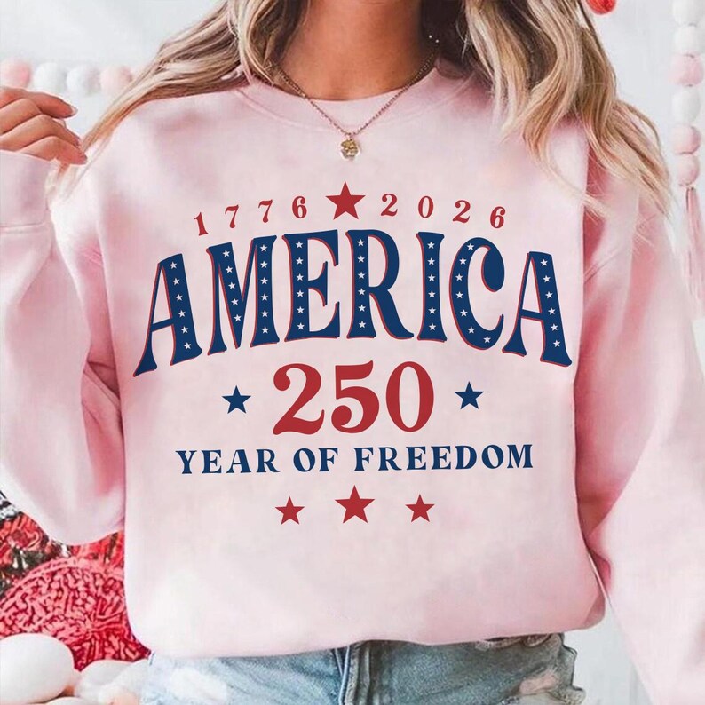 Puede incluir: Una sudadera rosa claro con el texto "1776 2026 AMERICA 250 YEAR OF FREEDOM" en rojo y azul. La palabra "AMERICA" est&aacute; en negrita, en arco con estrellas blancas. El n&uacute;mero "250" es grande y rojo.
