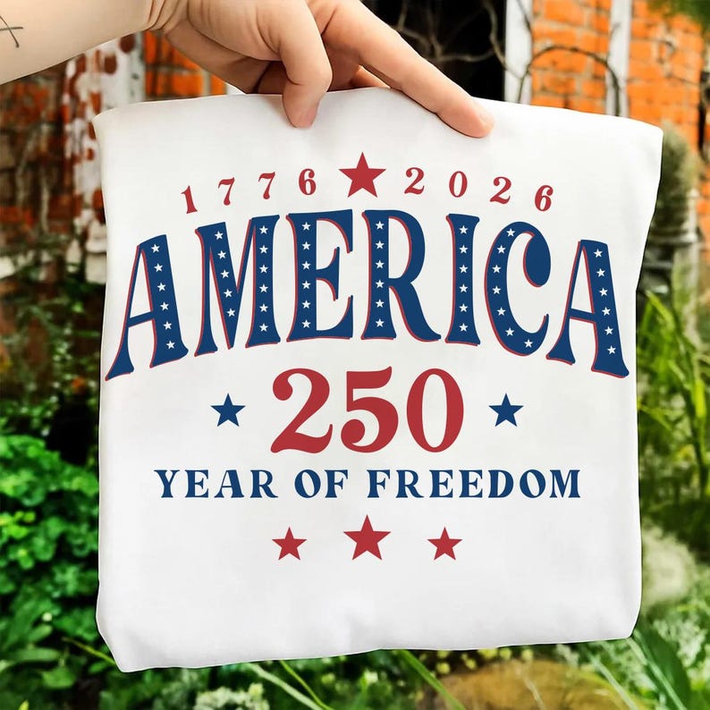 Puede incluir: Camiseta blanca con el texto "AMERICA 250 YEAR OF FREEDOM" en rojo y azul. El dise&ntilde;o incluye estrellas y los a&ntilde;os 1776 y 2026. La camiseta est&aacute; sujeta por una mano.