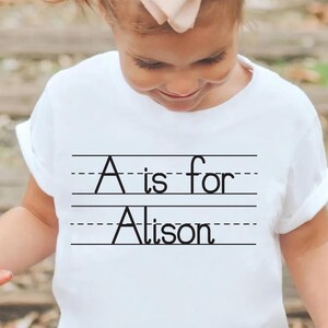 Puede incluir: Camiseta blanca con el texto negro "A is for Alison", escrito en un estilo de escritura escolar. El texto est&aacute; centrado en la camiseta, con l&iacute;neas arriba y abajo.