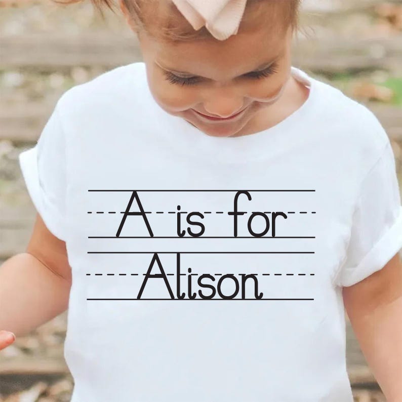 Puede incluir: Camiseta blanca con el texto negro "A is for Alison", escrito en un estilo de escritura escolar. El texto est&aacute; centrado en la camiseta, con l&iacute;neas arriba y abajo.