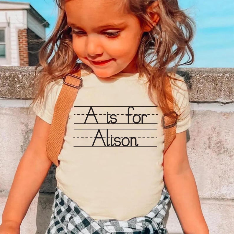 Puede incluir: Camiseta amarilla clara con el texto "A is for Alison" en negro. La camiseta la lleva un ni&ntilde;o que lleva una mochila marr&oacute;n. Ropa para ni&ntilde;os.