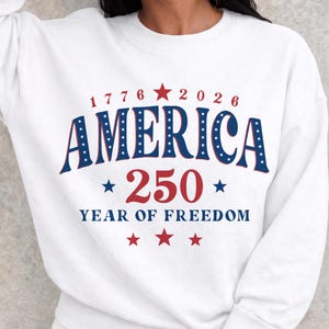 Puede incluir: Sudadera blanca con la palabra "AMERICA" en azul, delineada con estrellas blancas. El n&uacute;mero "250" es rojo, con el texto "YEAR OF FREEDOM" debajo. La sudadera tambi&eacute;n tiene los n&uacute;meros "1776" y "2026".
