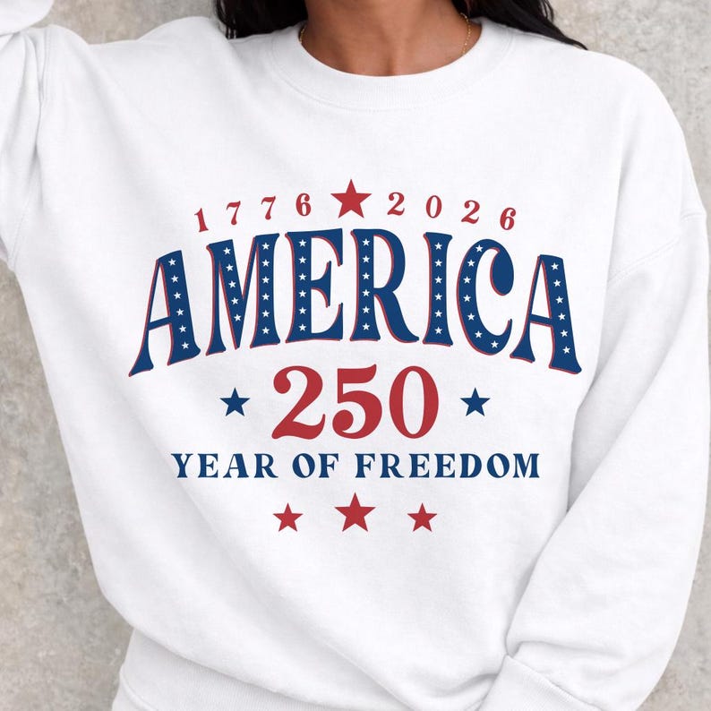 Puede incluir: Sudadera blanca con la palabra "AMERICA" en azul, delineada con estrellas blancas. El n&uacute;mero "250" es rojo, con el texto "YEAR OF FREEDOM" debajo. La sudadera tambi&eacute;n tiene los n&uacute;meros "1776" y "2026".