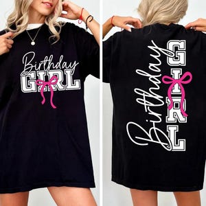 May include: Black t-shirt with the text "Birthday Girl" in white script and block letters, accented with a pink bow. The back of the shirt also features the text "Birthday Girl" in a vertical design with a pink bow.