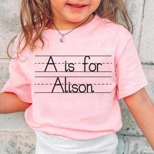 Puede incluir: Una camiseta rosa claro con el texto "A is for Alison" en negro, escrito sobre papel rayado. La camiseta de manga corta parece estar hecha de un material suave. La camiseta la lleva un ni&ntilde;o peque&ntilde;o.
