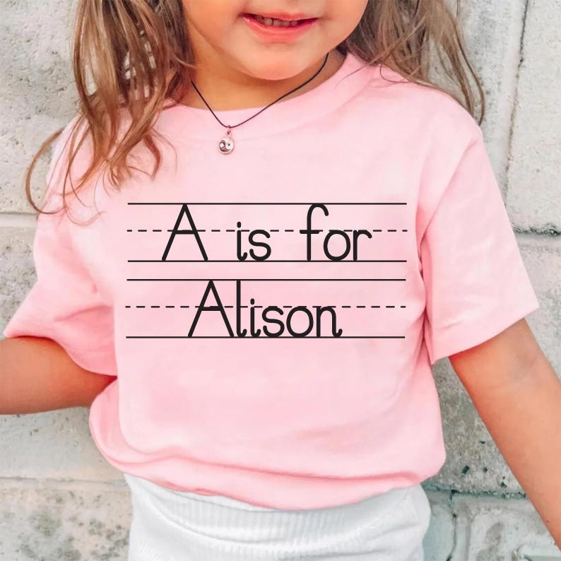 Puede incluir: Una camiseta rosa claro con el texto "A is for Alison" en negro, escrito sobre papel rayado. La camiseta de manga corta parece estar hecha de un material suave. La camiseta la lleva un ni&ntilde;o peque&ntilde;o.