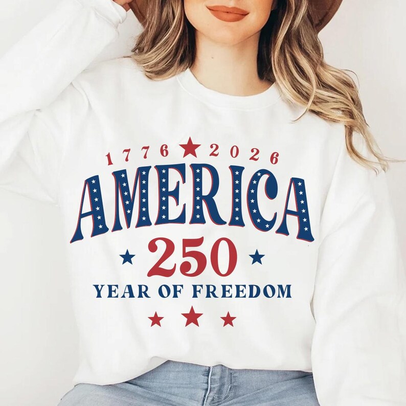 Puede incluir: Sudadera blanca con el texto "1776 2026 AMERICA 250 YEAR OF FREEDOM" en rojo y azul. La palabra "AMERICA" est&aacute; en letras azules grandes y en negrita con estrellas blancas. El n&uacute;mero "250" es rojo con estrellas a ambos lados.