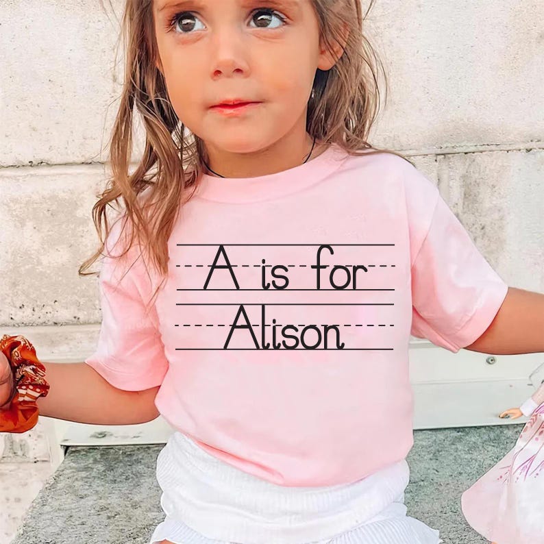 Puede incluir: Camiseta rosa claro con el texto negro "A is for Alison" sobre papel rayado. La camiseta es de manga corta.