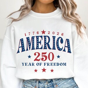 Puede incluir: Sudadera blanca con un dise&ntilde;o patri&oacute;tico. El dise&ntilde;o incluye la palabra "AMERICA" en azul con estrellas blancas, los n&uacute;meros "1776" y "2026", y "250 YEAR OF FREEDOM" en rojo y azul. Una prenda festiva.