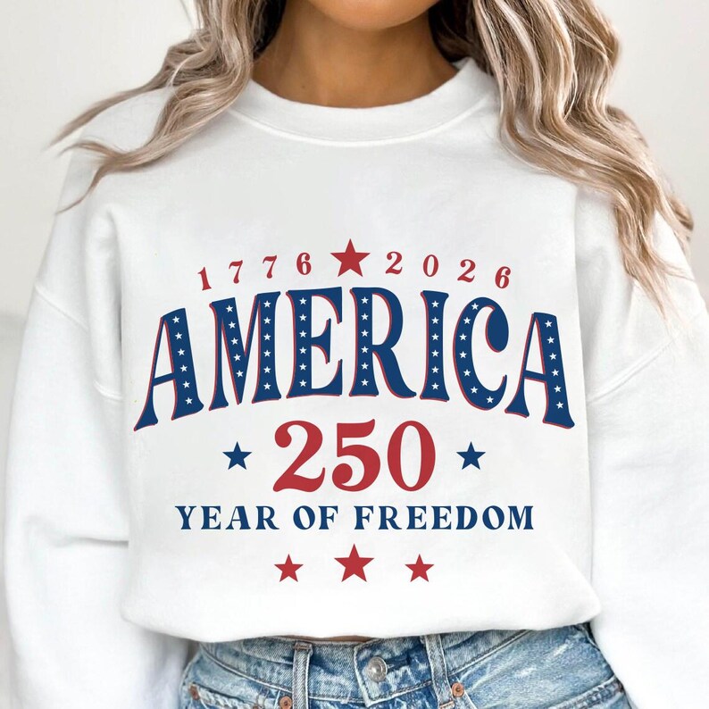 Puede incluir: Sudadera blanca con un dise&ntilde;o patri&oacute;tico. El dise&ntilde;o incluye la palabra "AMERICA" en azul con estrellas blancas, los n&uacute;meros "1776" y "2026", y "250 YEAR OF FREEDOM" en rojo y azul. Una prenda festiva.