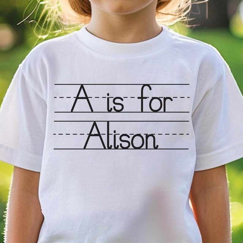Puede incluir: Camiseta blanca con el texto negro "A is for Alison", escrito en un estilo de escritura escolar. La camiseta tiene cuello redondo y mangas cortas. El fondo es verde borroso.