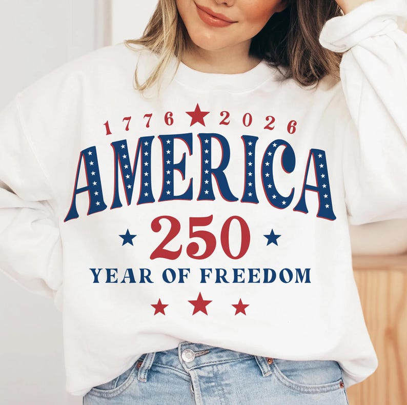 Puede incluir: Sudadera blanca con el texto "AMERICA 250 YEAR OF FREEDOM" en rojo y azul. El dise&ntilde;o incluye estrellas y los a&ntilde;os 1776 y 2026. Una prenda de vestir con tem&aacute;tica patri&oacute;tica.