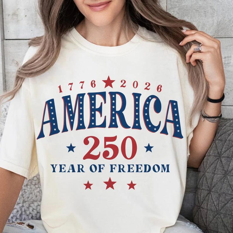 Puede incluir: Camiseta color crema con el texto "AMERICA 250 YEAR OF FREEDOM" en rojo y azul. El dise&ntilde;o incluye estrellas y los a&ntilde;os 1776 y 2026, celebrando el 250 aniversario.