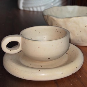 Op de afbeelding: Een crèmekleurige keramische kop en schotel set met een afgeronde handgreep en een gespikkelde afwerking. De kop staat op een bijpassende schotel, met een grotere kom op de achtergrond. De set is handgemaakt en heeft een rustieke, minimalistische uitstraling.