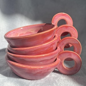 May include: Four coral-coloured ceramic bowls stacked, each with a distinctive ring handle. The bowls have a smooth, glossy finish. These handmade bowls are suitable for serving soups or small dishes. Each bowl is approximately 15 cm in diameter.