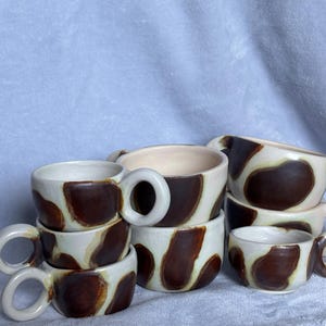 May include: A stack of handmade ceramic mugs with a unique cow-print design. The mugs are white with brown irregular patches, featuring rounded handles. The interior of the mugs is a creamy white color.