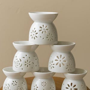 Peut inclure: Une pile de six diffuseurs d'huiles essentielles en céramique blanche. Chaque diffuseur a un dessus en forme de bol et une base avec des motifs floraux découpés. Ils sont disposés sur une surface en bois.