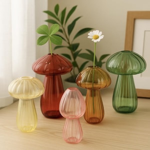 Peut inclure: Cinq vases en verre en forme de champignon, de couleurs jaune, rouge, orange, rose et vert. Chaque vase contient une seule fleur ou tige de plante, créant une présentation décorative. Les vases sont disposés sur une surface en bois.