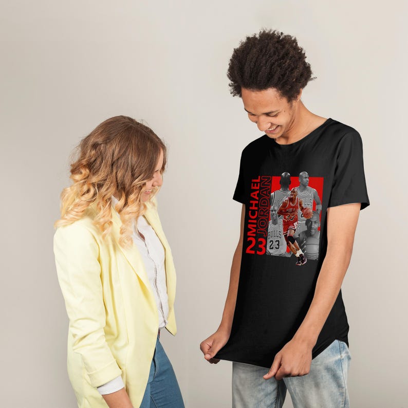 May include: Black t-shirt featuring a graphic of Michael Jordan in a red and white Chicago Bulls uniform. The text "MICHAEL JORDAN 23" is printed vertically in red. The shirt is being held up by a person.