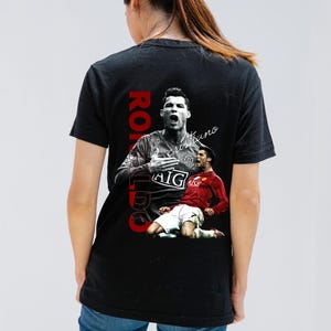 Pu&ograve; includere: T-shirt nera con grafica di un calciatore in maglia rossa e bianca. La parola "RONALDO" &egrave; stampata verticalmente in rosso, con il nome "cristiano" in corsivo.