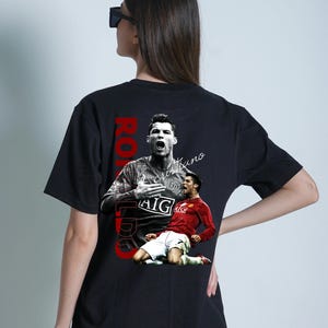 Pu&ograve; includere: T-shirt nero con grafica di un calciatore in maglia rossa, con il nome "Ronaldo" in testo verticale rosso. La maglietta include anche il testo "Cristiano".