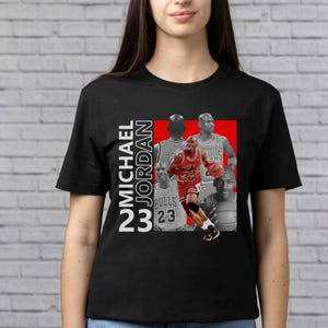 May include: Black t-shirt featuring a graphic of Michael Jordan in a red and white design. The design includes the text "MICHAEL JORDAN 23" in white. The shirt has a crew neck and short sleeves.