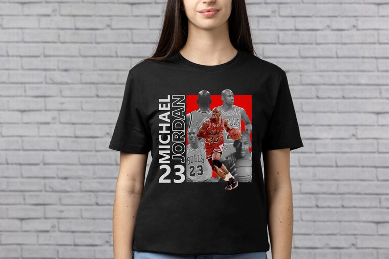 May include: Black t-shirt featuring a graphic of Michael Jordan in a red and white design. The design includes the text "MICHAEL JORDAN 23" in white. The shirt has a crew neck and short sleeves.